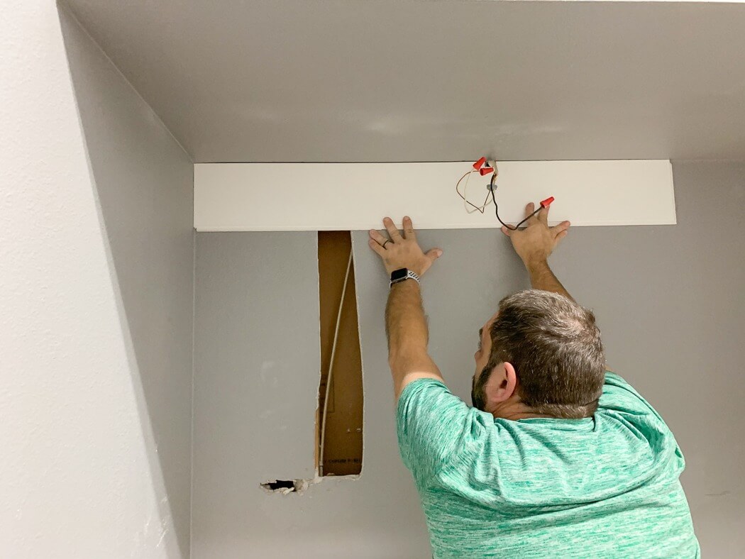 A man installing a white panel on a wall with exposed wires, wearing a green shirt and working in a small alcove.