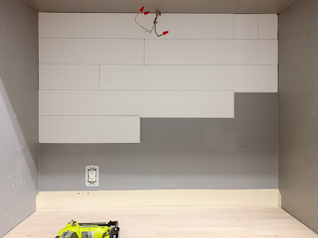 Partially installed white shiplap on a gray wall with exposed wires, a power tool on the wooden countertop below.