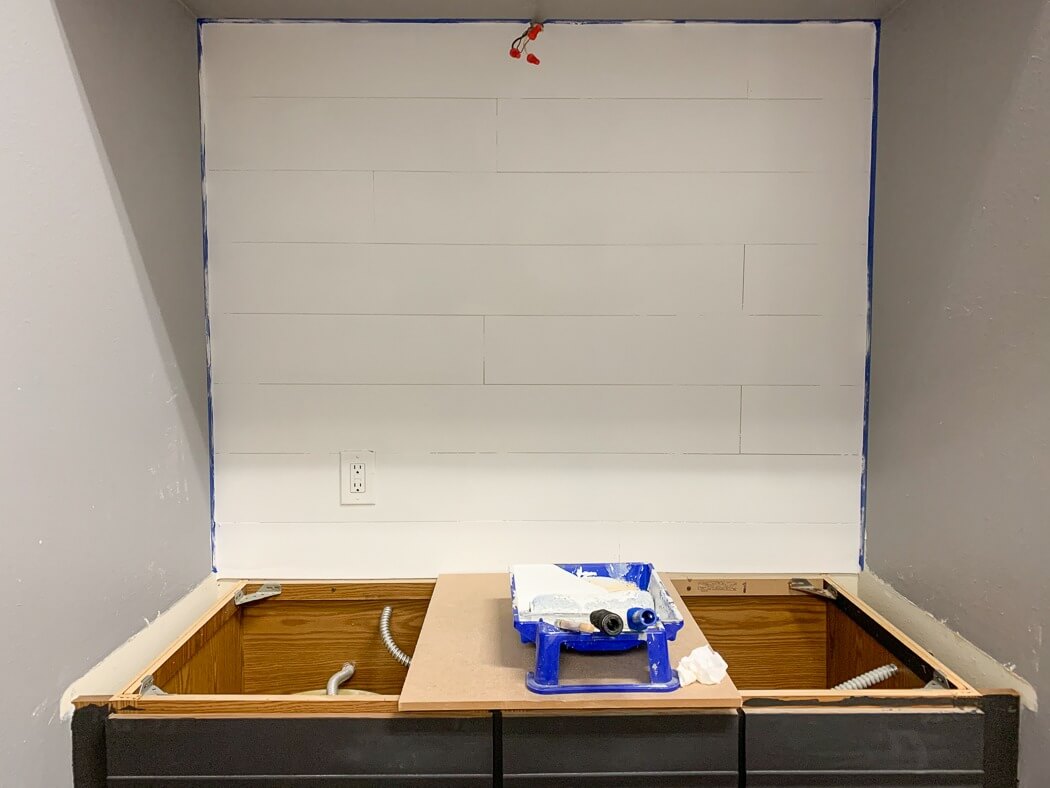 Partially painted shiplap wall with blue painter's tape along the edges, and a paint tray with rollers on a countertop under construction.