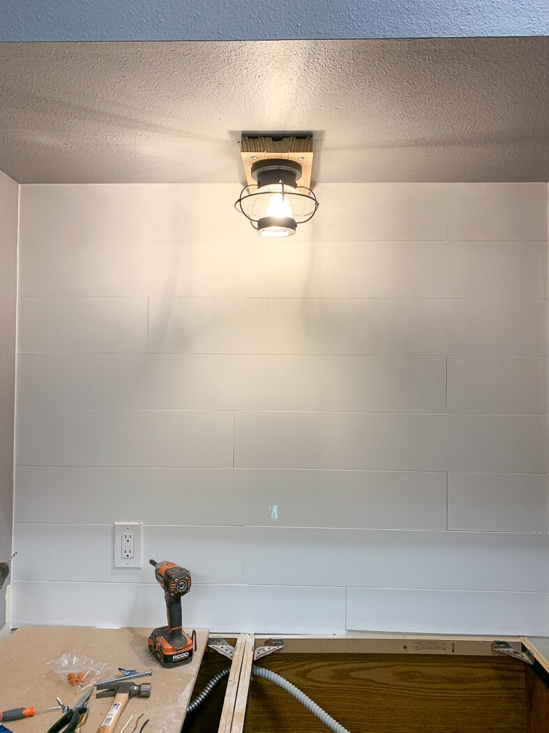 Newly installed light fixture illuminating a shiplap wall with tools and a drill on a partially assembled countertop below.
