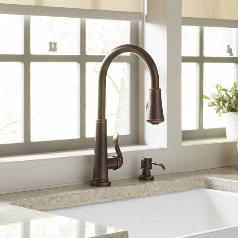 Close-up of a bronze kitchen faucet with a soap dispenser, set on a light-colored granite countertop in front of large windows with a small potted plant in the background.