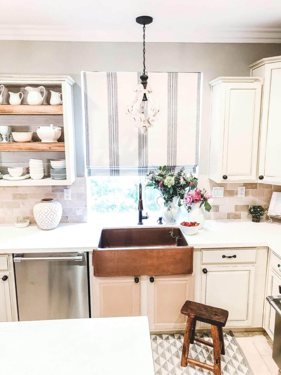 Charming kitchen with a hammered copper farmhouse sink, cream-colored cabinets, a patterned rug, and open shelving displaying white dishware.
