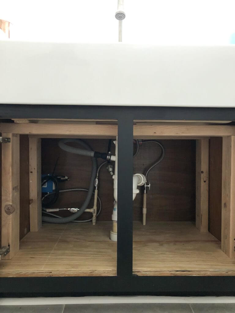 The plumbing setup underneath a sink, showing exposed pipes and fittings within a wooden cabinet frame.
