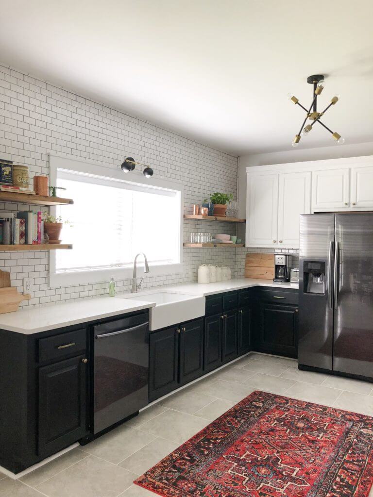 A modern kitchen with black lower cabinets, white upper cabinets, a large farmhouse sink, stainless steel appliances, and a subway tile backsplash, featuring a red patterned rug on the floor.