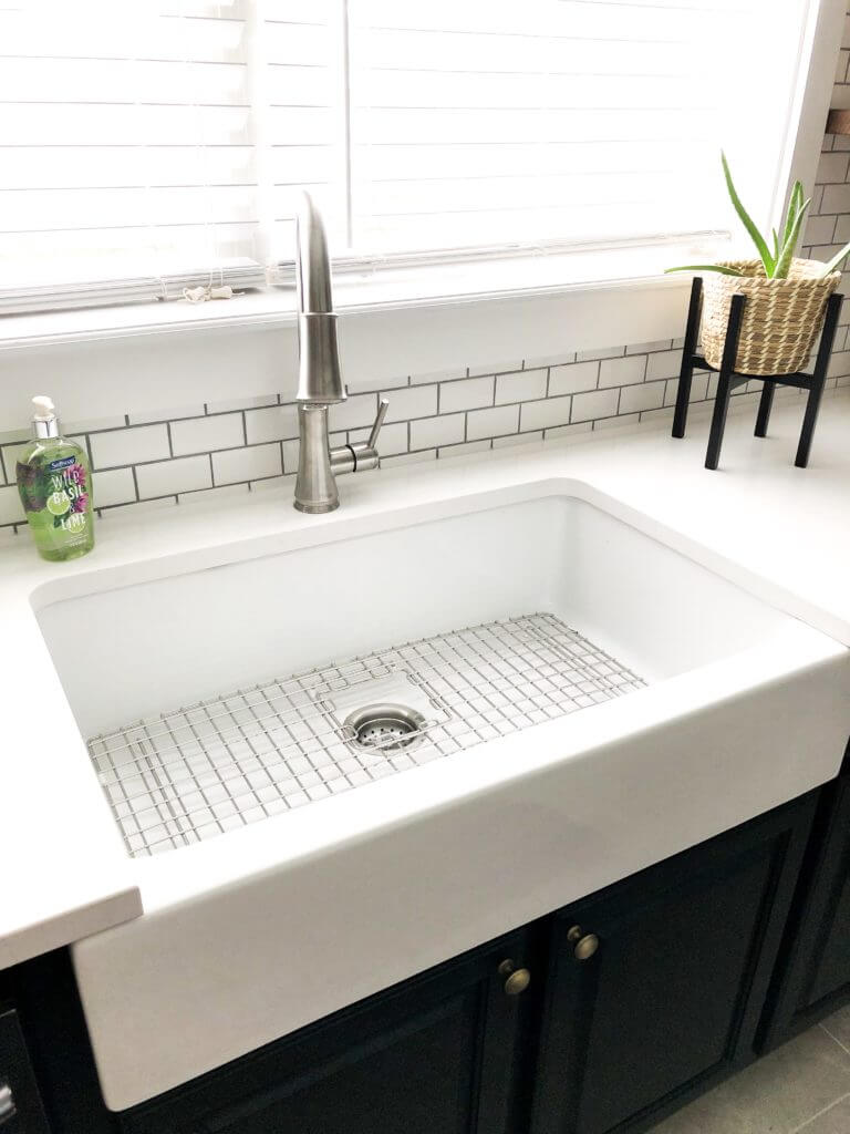 A bright kitchen sink area with a large white farmhouse sink, stainless steel faucet, subway tile backsplash, and an aloe plant in a woven basket.