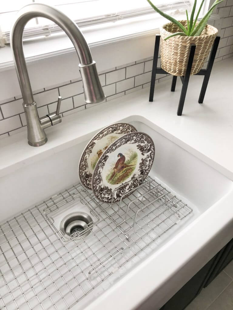 A white farmhouse sink with a stainless steel faucet, featuring decorative plates in a drying rack and an aloe plant in a woven basket on the countertop.