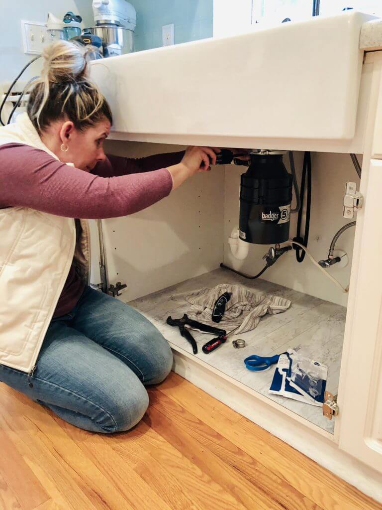 A woman kneeling on the floor, working under a kitchen sink with tools and a garbage disposal unit visible.