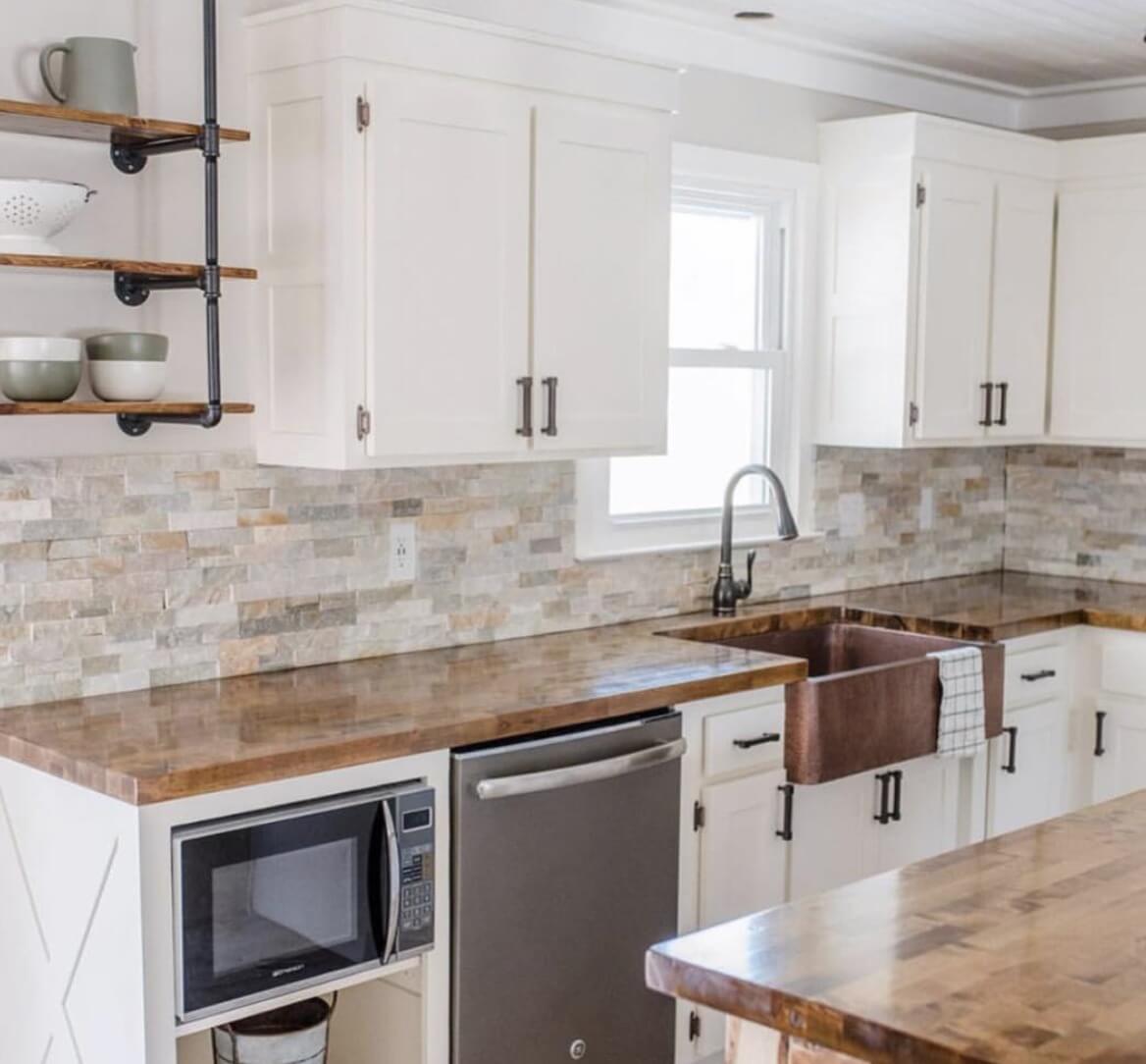 Bright kitchen with white cabinets, wooden countertops, a copper farmhouse sink, and a stone tile backsplash, featuring open shelves with dishes and a stainless steel dishwasher.