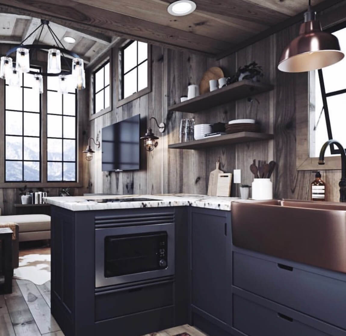 A cozy rustic kitchen featuring dark cabinetry, a copper sink, marble countertops, and wooden walls with large windows overlooking a mountain view. The space is accented with a copper pendant light, open shelving, and a modern chandelier.