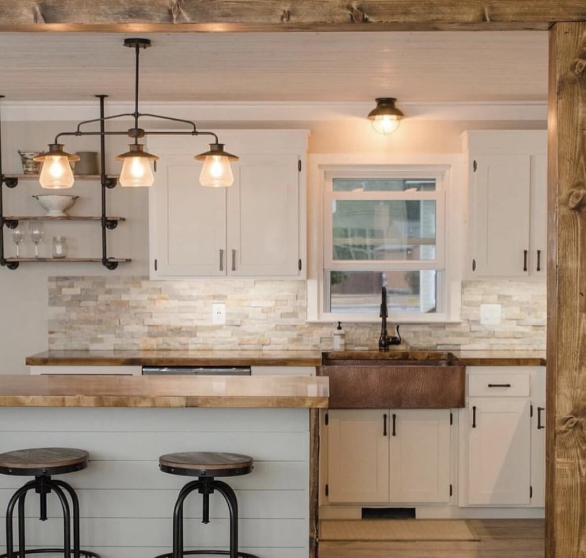 A warm farmhouse-style kitchen with white cabinetry, a copper sink, wooden countertops, and a stone backsplash. The space features industrial-style pendant lights and open shelving, creating a cozy and inviting atmosphere.