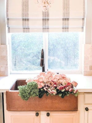Bright kitchen scene with a large copper farmhouse sink filled with pink and green flowers, under a window with striped Roman shades. Cream-colored cabinets and light countertops add to the airy feel.