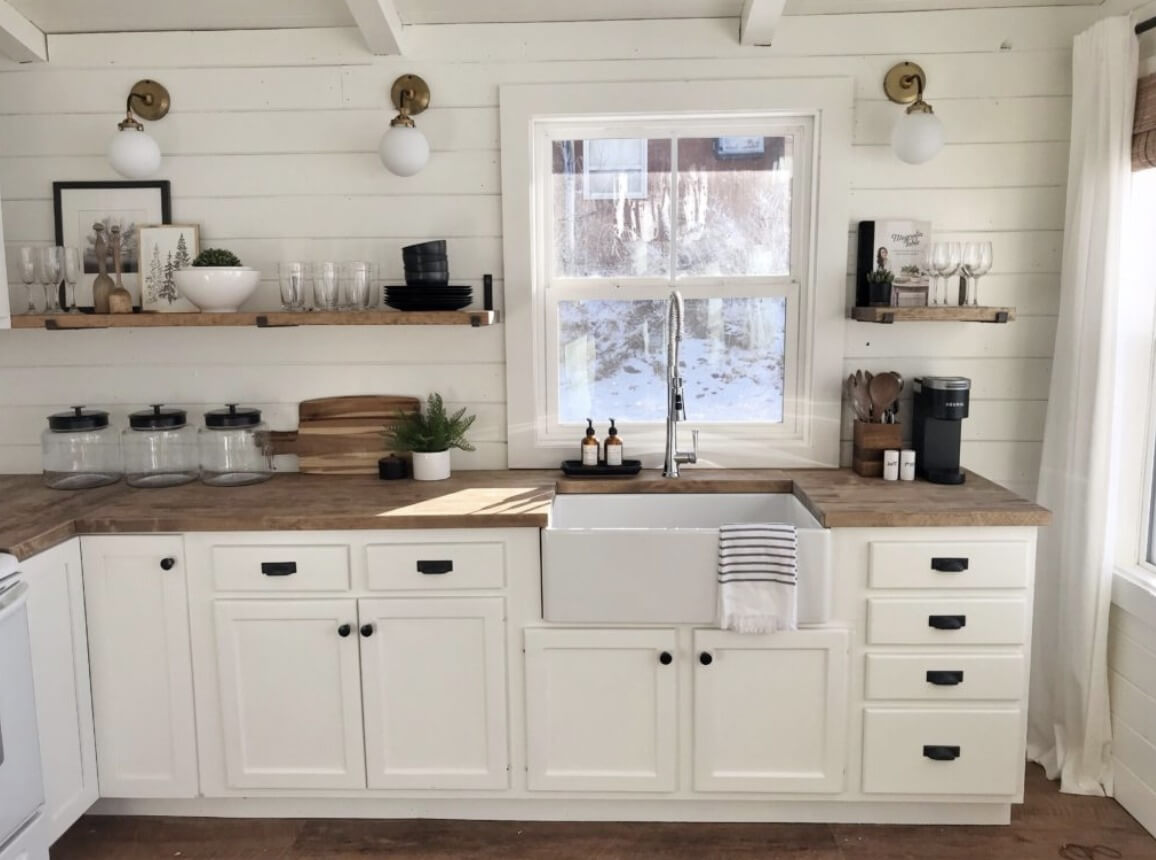A bright kitchen with white cabinets, a farmhouse sink, and wooden countertops. Shelves hold dishes, decor, and a coffee maker, while large windows provide natural light. Two wall-mounted lights are above the shelves.