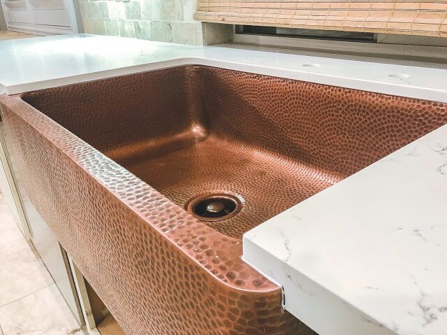 Close-up of a hammered copper farmhouse sink set into a white marble countertop, with a textured backsplash and bamboo blinds in the background.