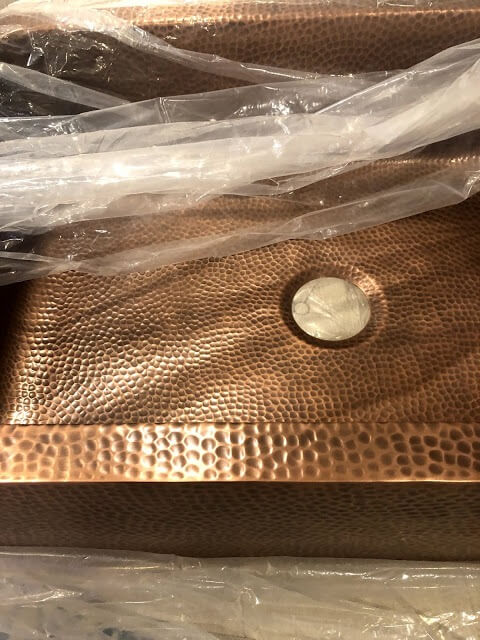 Close-up of a hammered copper sink, partially wrapped in protective plastic, highlighting its rich texture and warm metallic finish.