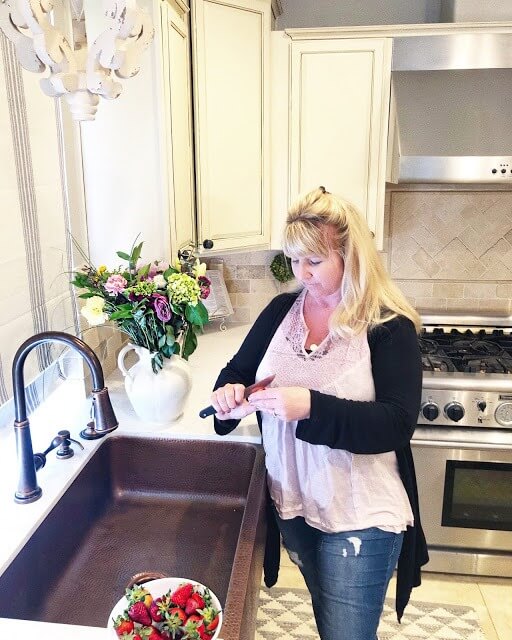 A woman standing in a bright kitchen beside a copper farmhouse sink, preparing strawberries, with a bouquet of flowers on the counter and a stainless steel stove in the background.