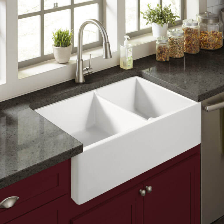 A bright kitchen featuring a white double-basin farmhouse sink with a dark granite countertop, stainless steel faucet, and red cabinetry, set against a backdrop of large windows with potted plants and jars on the windowsill.