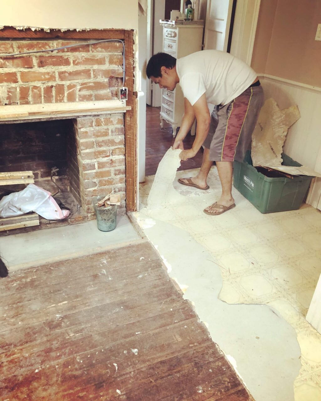 A man working on a home renovation project, removing old flooring near an exposed brick fireplace, with tools and debris scattered around.