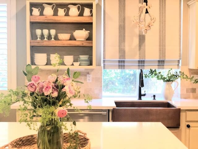 A bright and airy kitchen featuring a copper farmhouse sink beneath a window with striped Roman shades, open shelving displaying white dishware, and a vase of pink flowers on the countertop.