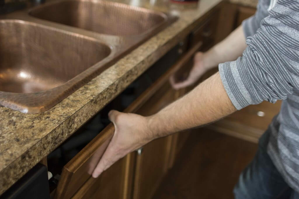 A person is removing the front panel of a kitchen cabinet beneath a double copper sink.