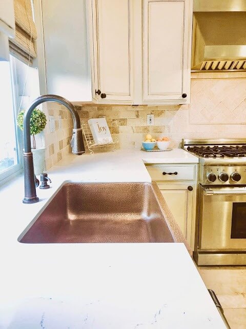 Bright kitchen with a hammered copper sink, bronze faucet, cream-colored cabinets, and a stainless steel stove. The countertop is decorated with a small potted plant and a sign that reads "Joy."