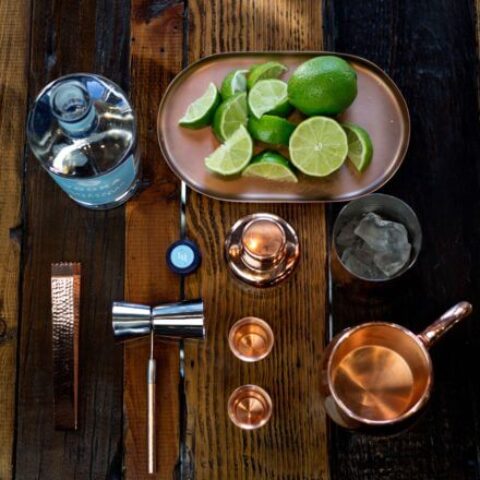 A wooden table set with ingredients and tools for making cocktails, including a bottle, sliced limes, a copper mug, and shot glasses.