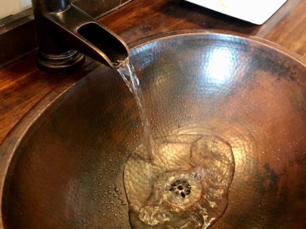 A close-up of a copper sink with water flowing from a faucet, highlighting the sink's hammered texture and rich, warm tones against a wooden countertop.