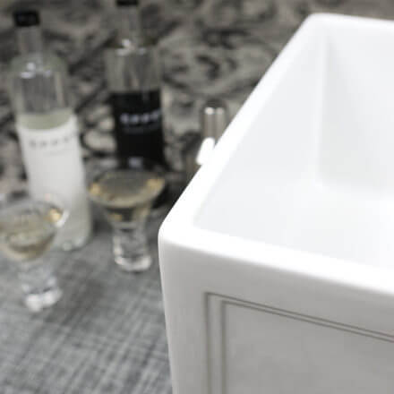 Close-up of a white farmhouse sink with blurred bottles and glasses in the background, suggesting a kitchen or bar setting.