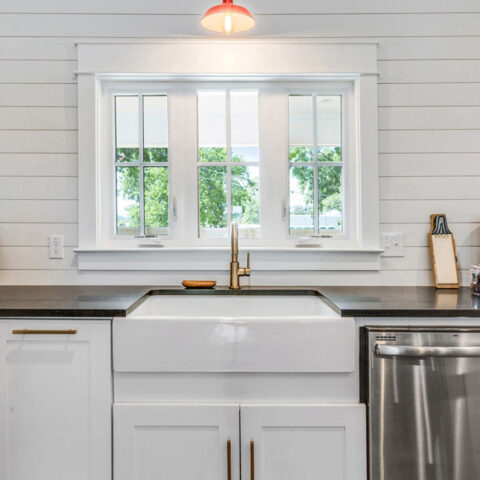 A farmhouse-style kitchen features a white apron-front sink with a gold faucet, set against a black countertop. A window with a view of greenery is positioned above the sink, and a red pendant light hangs overhead. Stainless steel appliances and simple decor complete the space.