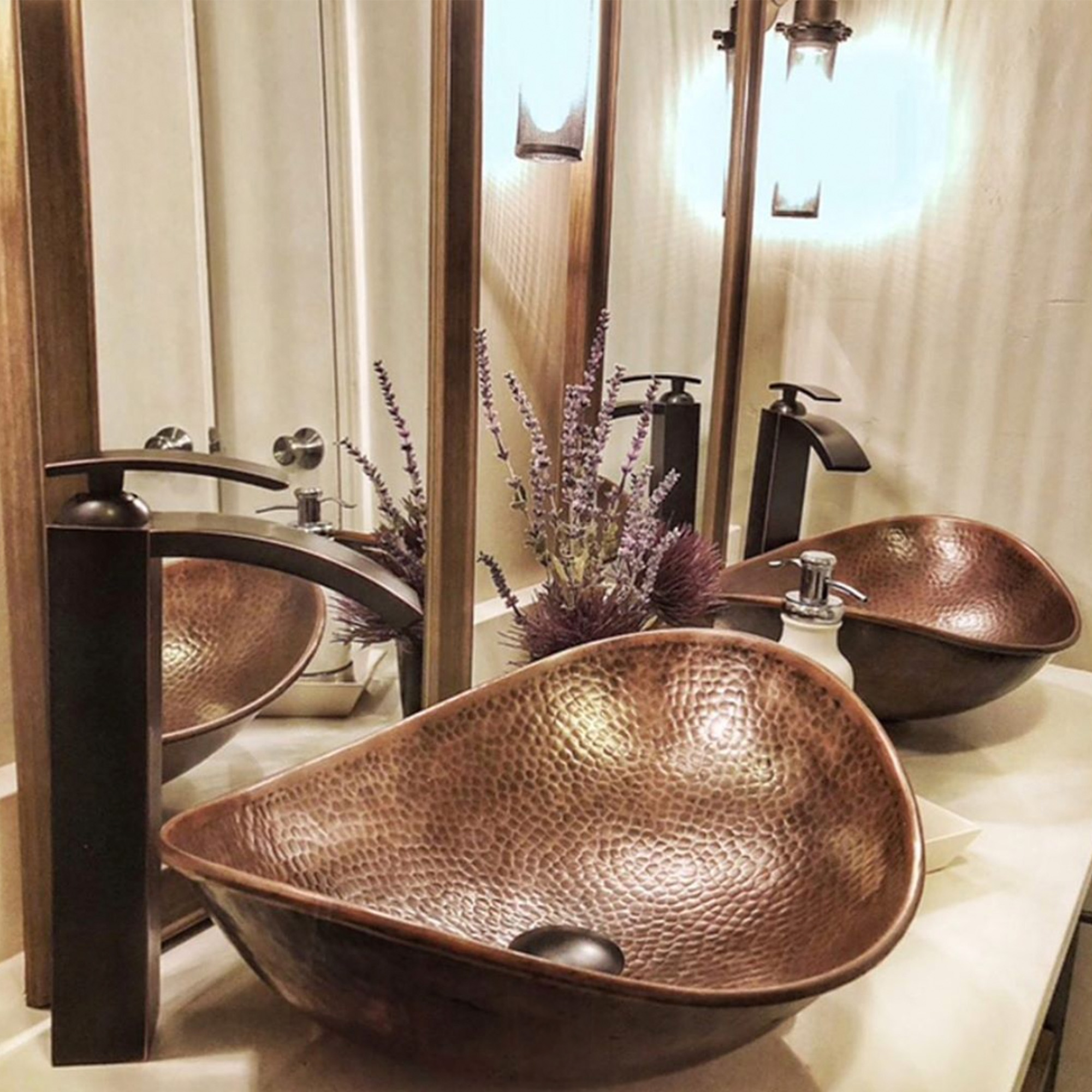 A duo of hand hammered copper vessel bathroom sinks with antique brown faucets in a bathroom.