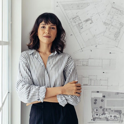 Professionally dressed female architect leaning against an office wall full of building plans and designs
