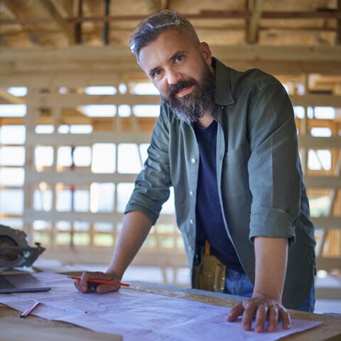 Male general contractor standing at a table of architectural plans inside of a room with unfinished walls and exposed wood framing.