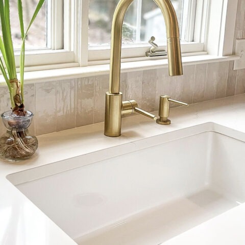 Fireclay white kitchen sink with gold faucet under a window with white countertop