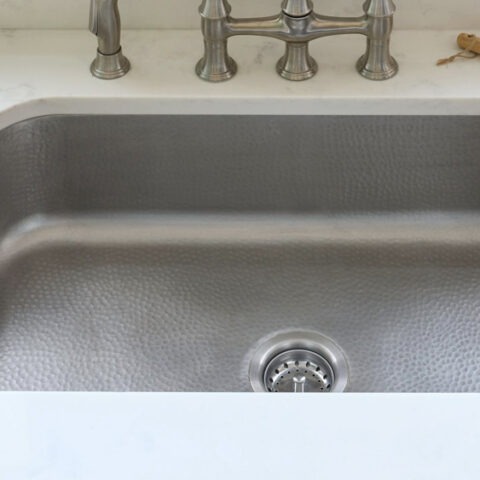 Undermount hammered stainless steel kitchen sink with floral wallpaper backdrop and vintage bridge faucet