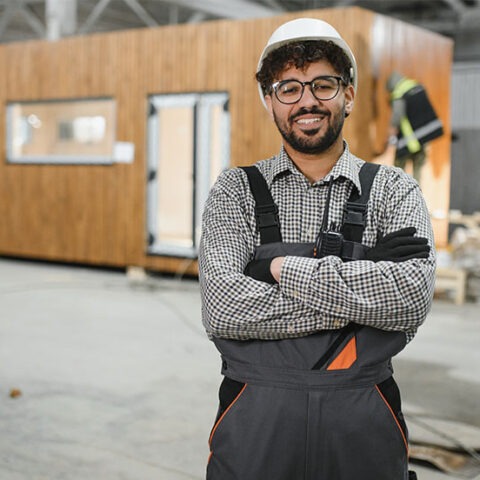A male wearing overalls and hardhat in a warehouse of manufactured tiny homes